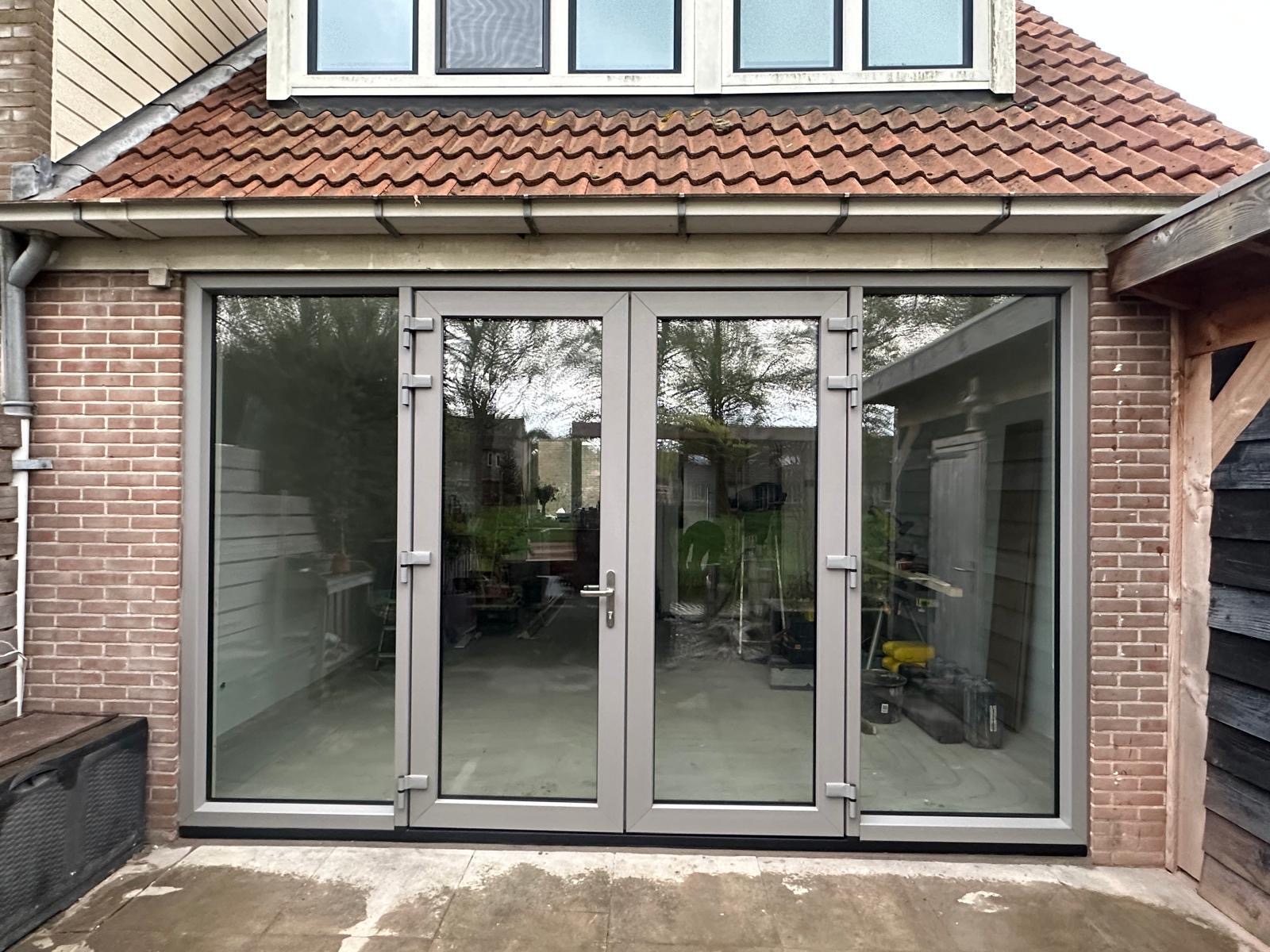 Grijze kunststof tuindeuren met twee openslaande vleugels en vaste zijlichten, gezien vanuit de tuin onder een pannendak